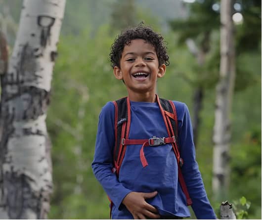 Barn klädd i en blå långärmad tröja och en rödkantad ryggsäck medan det står i en skog.