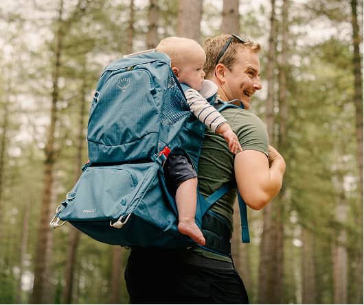 En vuxen som bär ett spädbarn i en blå Osprey-bärryggsäck för barn medan han går genom en skog.