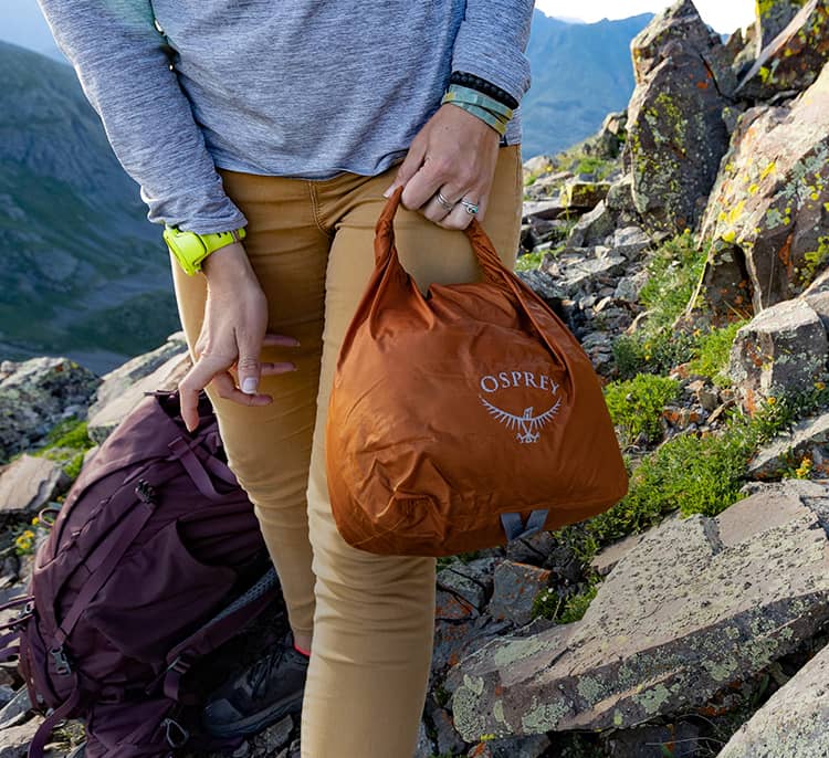 Person standing on a rocky mountainside holding a compact orange stuff sack, with hiking gear and terrain visible around them.