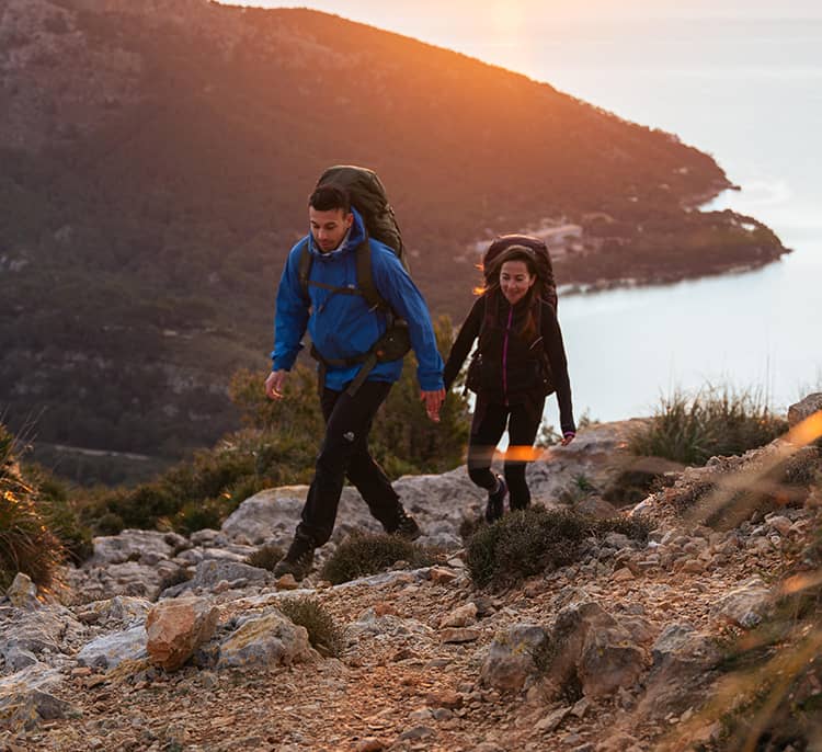 Two backpackers hike uphill on a rocky coastal trail at sunset, overlooking the ocean and surrounding hills.