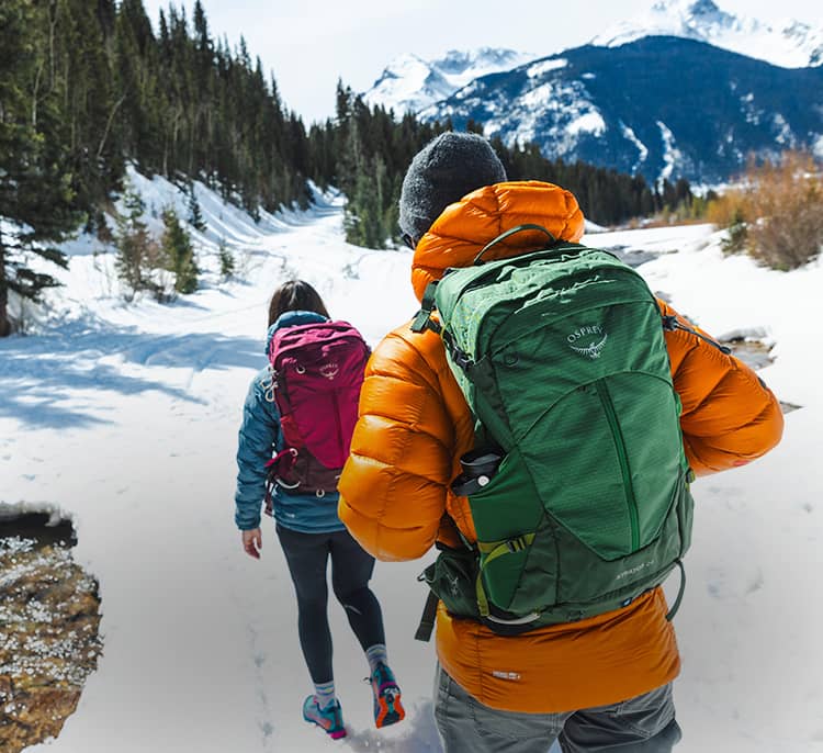 Two hikers walk along a snow-covered path in the mountains, wearing insulated jackets and carrying Stratos and Sirrus hiking backpacks with alpine scenery in the background.