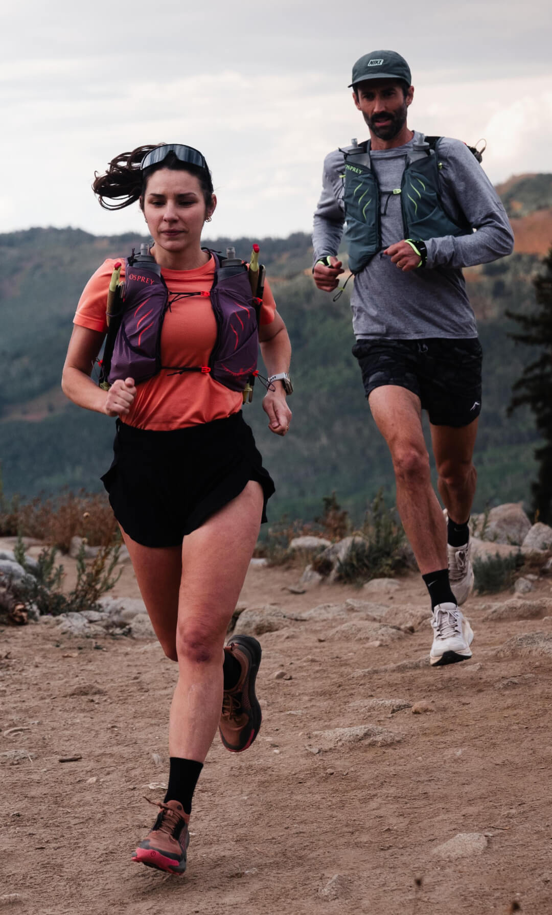 Due corridori da trail con giubbotti da corsa Osprey che corrono su un sentiero di montagna rocciosa.