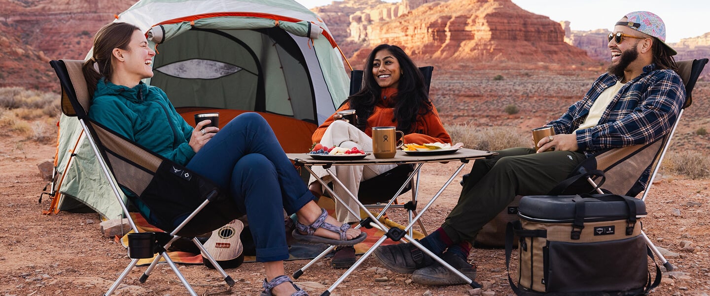 Group of campers sitting in foldable chairs around a camp table outside a tent in a desert landscape, using Osprey outdoor gear and travel accessories for camping and adventure.