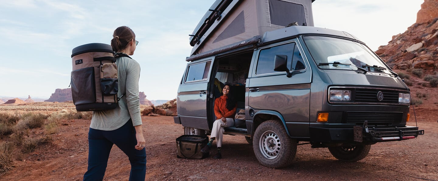 Person walking toward a camper van with rooftop tent in a desert landscape, carrying a large Osprey travel backpack, showcasing durable outdoor gear for overlanding and adventure travel.