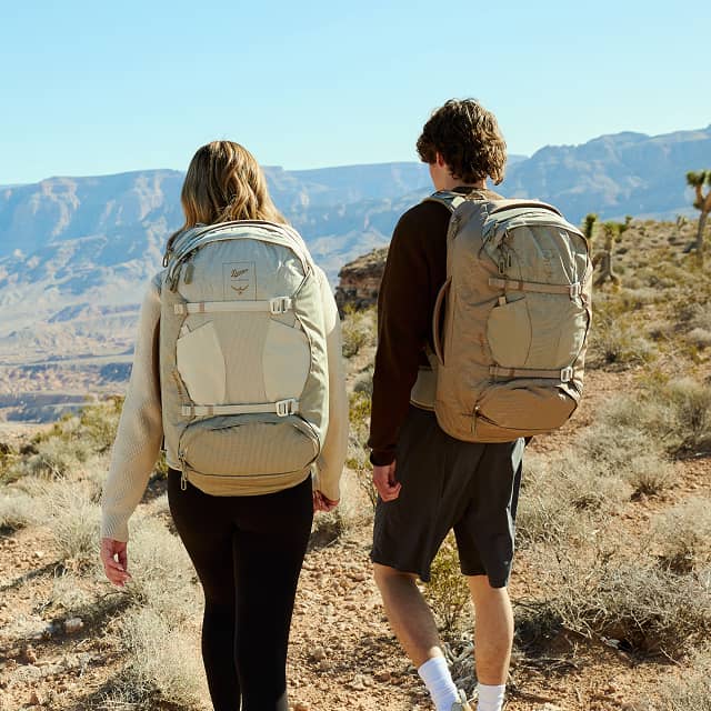 Two hikers wearing Osprey hiking backpacks walk along a desert trail overlooking a canyon landscape on an outdoor adventure.