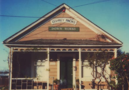 Verblichenes, altes Foto eines pfirsichfarbenen Hauses im Craftsman-Stil mit einer Veranda, die zu einem Laden mit zwei Schildern mit der Aufschrift „Osprey Packs“ und „Down Works“ umgebaut wurde.
