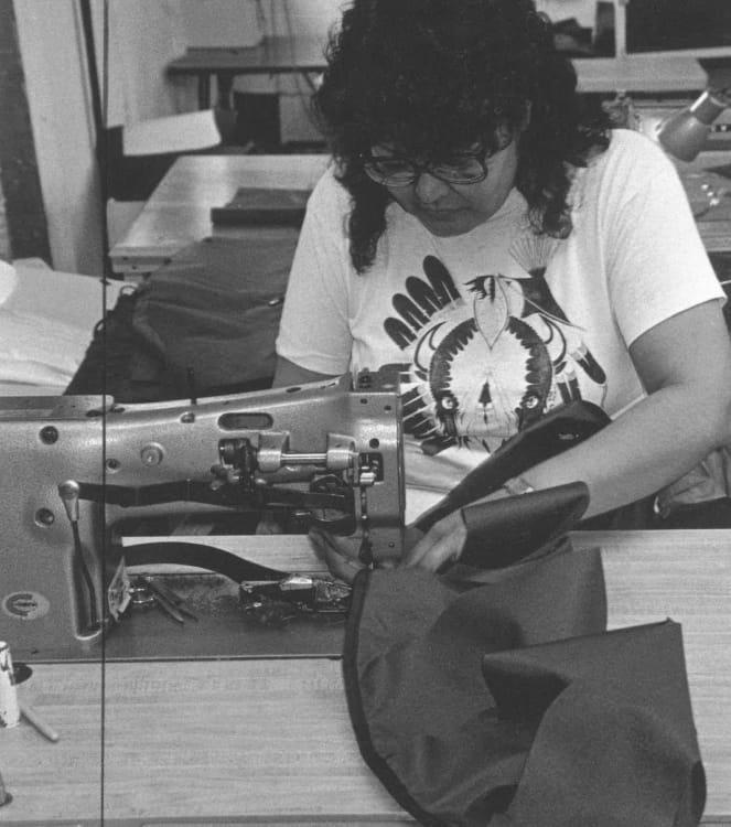 Schwarz-Weiß-Foto einer Frau mit lockigem schwarzem Haar, Brille und weißem Grafik-T-Shirt, die sich auf das Nähen von Rucksack-Bestandteilen mit einer Maschine konzentriert.