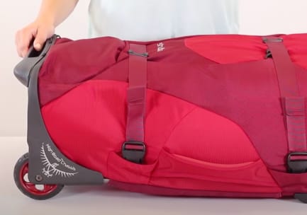 Eine Tasche mit roten Rollen liegt auf dem Rücken. Zwei StraightJacket Compression-Gurte mit gepolsterten Seitenwänden werden fest über das Hauptmaterial geschnallt. Der Rahmen der Rolle trägt ein Osprey-Abzeichen und die Aufschrift „High Road Chassis“. Eine Person in einem weißen T-Shirt hält das Gestell fest.