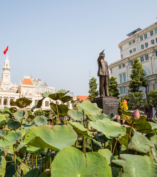 Lotusblätter und Knospen im Vordergrund und die Ho-Chi-Minh-Statue vor dem Rathaus von Saigon in Ho-Chi-Minh-Stadt, Vietnam.