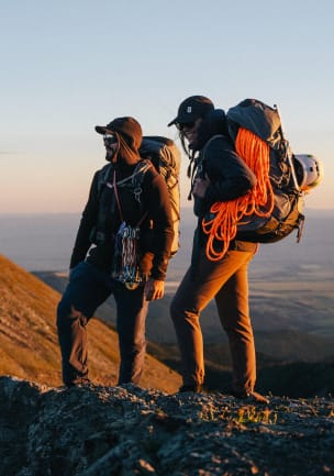 Zwei Kletterer stehen im Morgenlicht auf einem Bergkamm und tragen voll beladene Rucksäcke mit Kletterseil, Karabinern, Helmen und Eispickel.