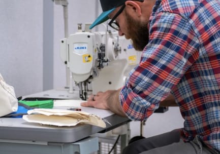 Ein bärtiger Mann mit Brille, Baseballmütze und einem blau-weiß-roten Flanellhemd mit hochgekrempelten Ärmeln fertigt an einer Nähmaschine eine Gesichtsmaske aus Stoff.