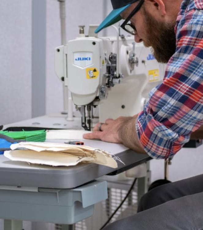 Ein bärtiger Mann mit Brille, Baseballmütze und einem blau-weiß-roten Flanellhemd mit hochgekrempelten Ärmeln fertigt an einer Nähmaschine eine Gesichtsmaske aus Stoff.