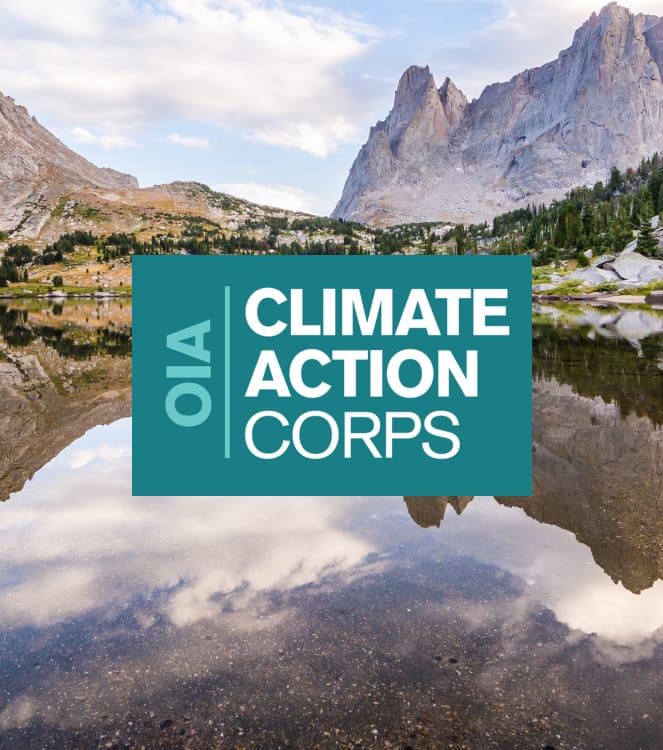 Das blaugrüne und weiße Logo des OIA Climate Action Corps über dem Foto einer bewaldeten Berglandschaft mit einem glasklaren, spiegelnden Bergsee und einem blauen Himmel.