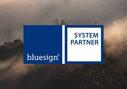 Das dunkelblaue und weiße bluesign SYSTEM PARTNER Logo über dem Foto eines nebligen, bewaldeten Berges.