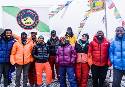 Ein Team von sieben Bergsteigern in Bergsteigerhosen und dicken Mänteln unter einer weiß-grün gestreiften Flagge mit blauen, roten und gelben Abzeichen, auf denen „Full Circle Everest Expedition 2022“ steht, und tibetischen Gebetsfahnen, die hinter ihnen im Wind wehen.