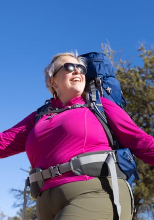 Ein Foto von einer Frau mit silbernen Haaren, die einen Fuchsia-farbenen Pullover mit halbem Reißverschluss, eine Wanderhose und einen kobaltblauen und grauen Osprey-Rucksack unter einem blauen Himmel trägt.