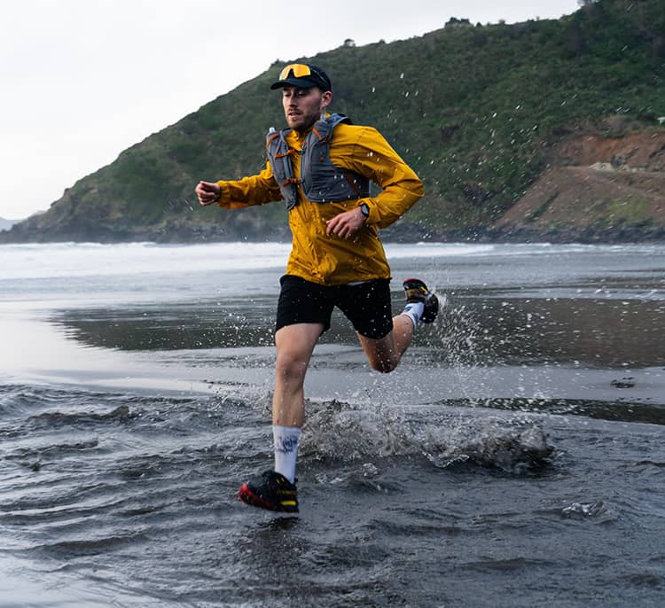 Runner splashes through shallow ocean water along a beach, wearing a lightweight jacket and Duro hydration vest with coastal hills in the background.