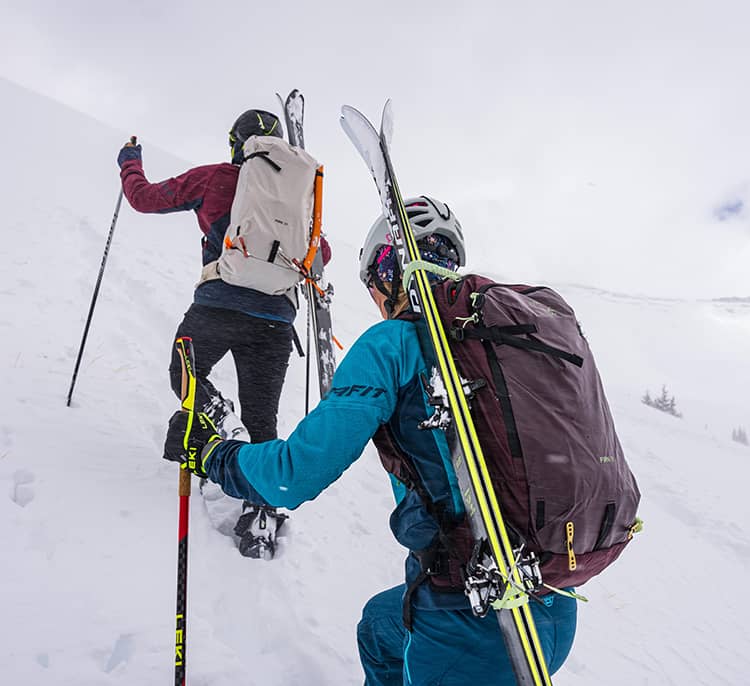 Two winter mountaineers climb a steep, snow‑covered slope using poles, carrying skis on their Osprey Firn backpacks in a high‑alpine environment.