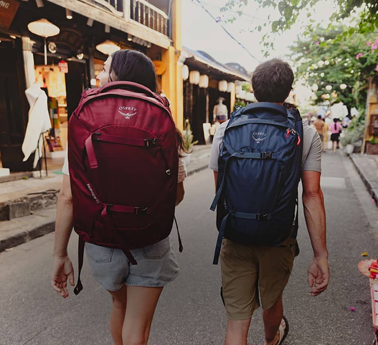 Two travelers walk down a narrow street lined with shops, each carrying a large travel Farpoint Fairview backpack while exploring an urban destination.
