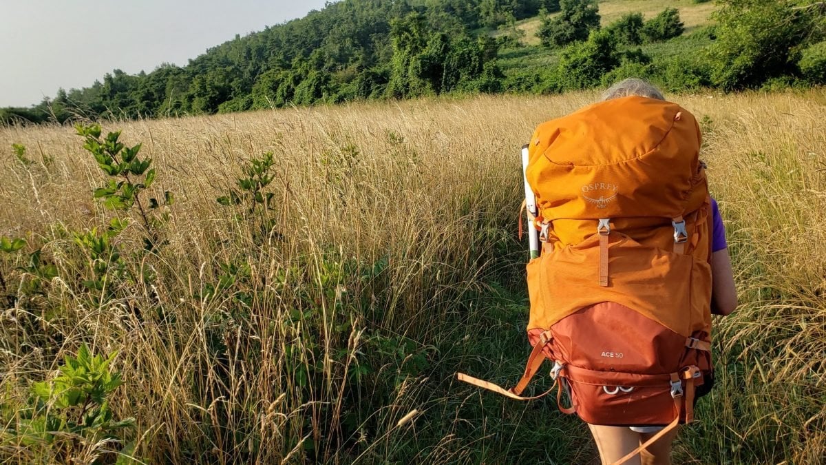 Ayla Crawley hiking through a field on the AT in her orange Opsrey Ace 50 pack
