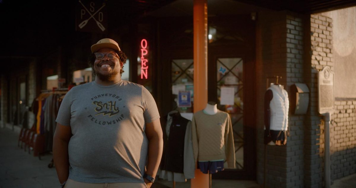 A man smiling and standing in front of his store, with clothing display racks behind him