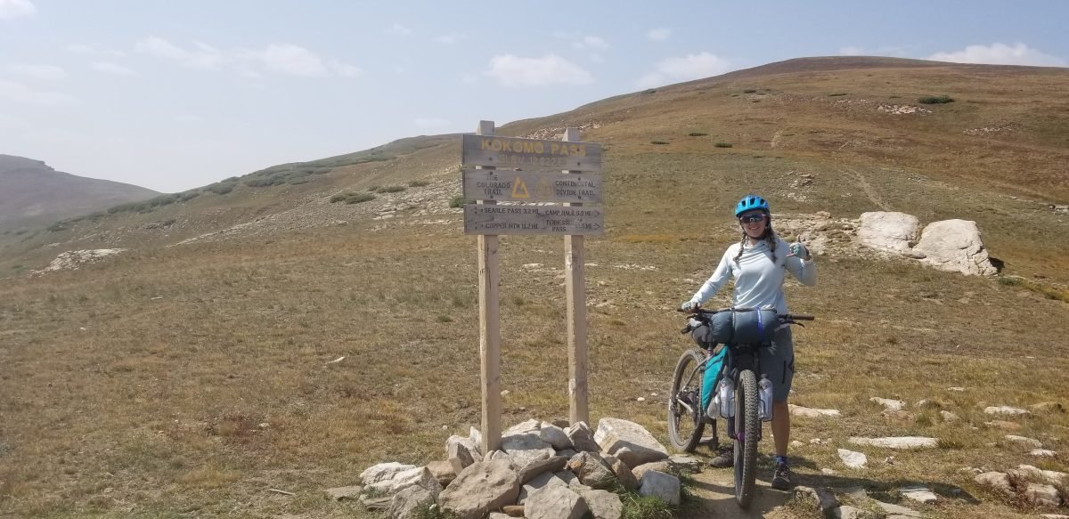 A woman standing next to a sign that reads Kokomo Pass holding her mountain bike next to her, which is packed with gear