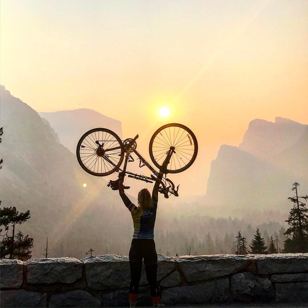 A person holding a bike upside down above their head, looking out onto a mountainous view of a sunset