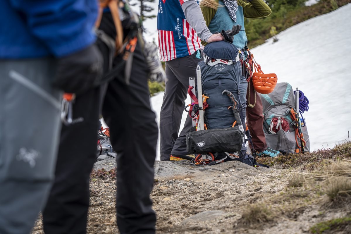 A close up of backpacking packs on the ground, with people surrounding