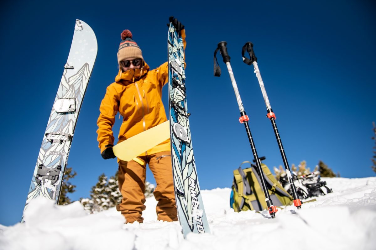 A woman wearing mustard yellow snow gear holding splitboarding gear in the snow