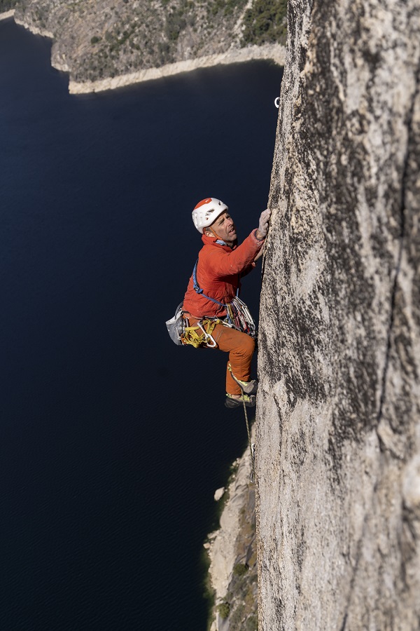 A man rock climbing up a cliff face, wearing a red top and orange pants, and a white and red helmet