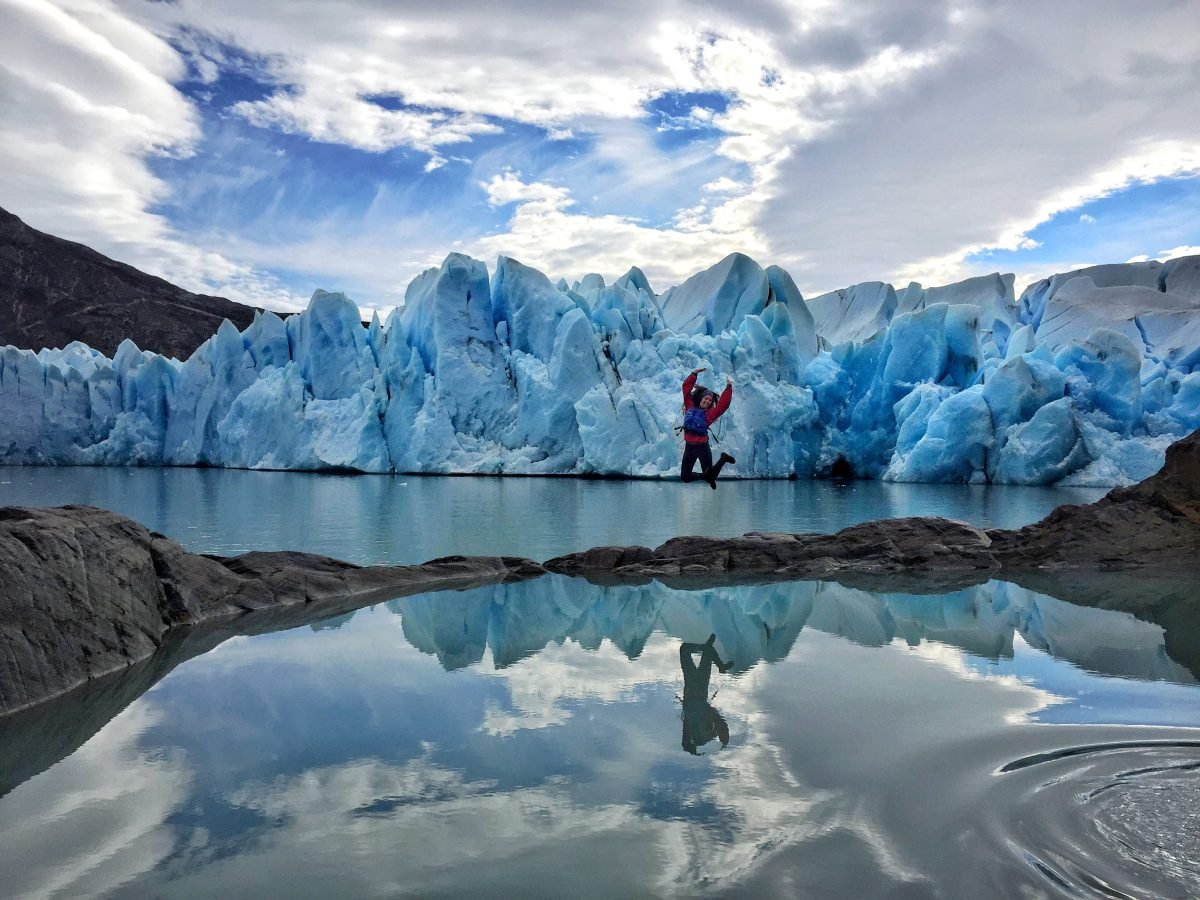 A woman wearing a red shirt and a blue life jacket jumps on a small path of rocks between two bodies of water, with glaciers in the background