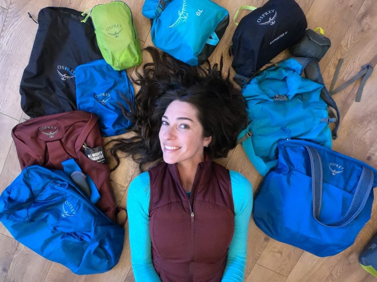A woman wearing a teal shirt and a maroon vest laying on the ground surrounded by travel accessories