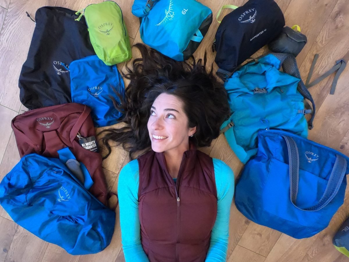 A woman wearing a teal shirt and a maroon vest laying on the floor, with travel gear surrounding her on all sides