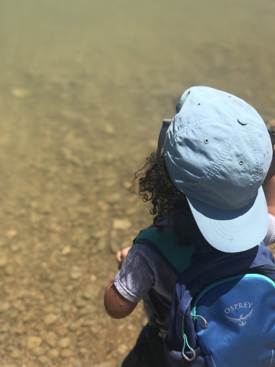 A young kid with long curly hair and a backwards hat wearing a blue backpack standing at waters edge