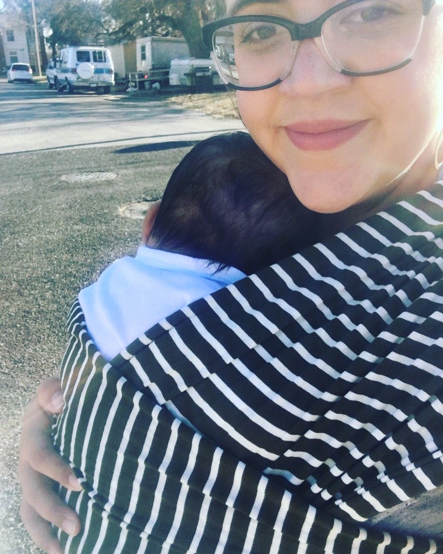 A woman walking outside with a baby cradled to her chest