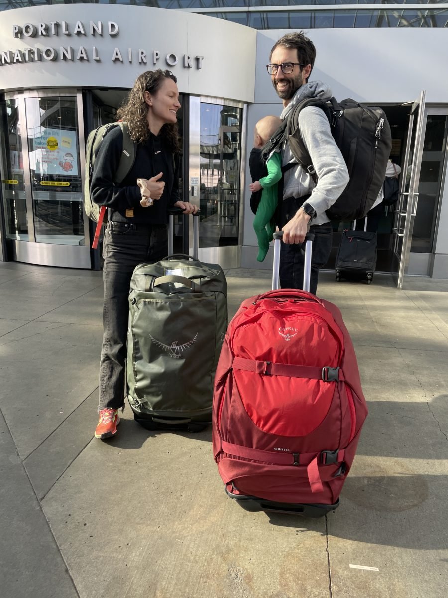 A man and woman standing in front of the Portland airport with their luggage, and their tiny baby
