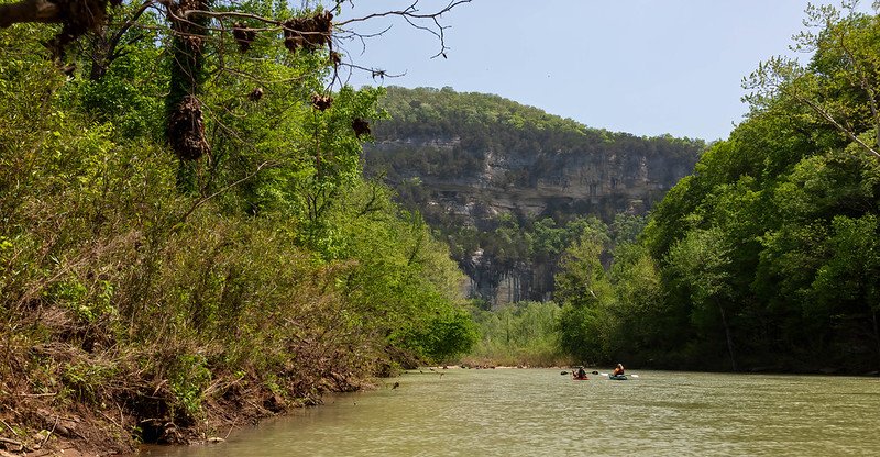 Two paddlers off in the distance on a river, with bluffs in the background and trees on either side
