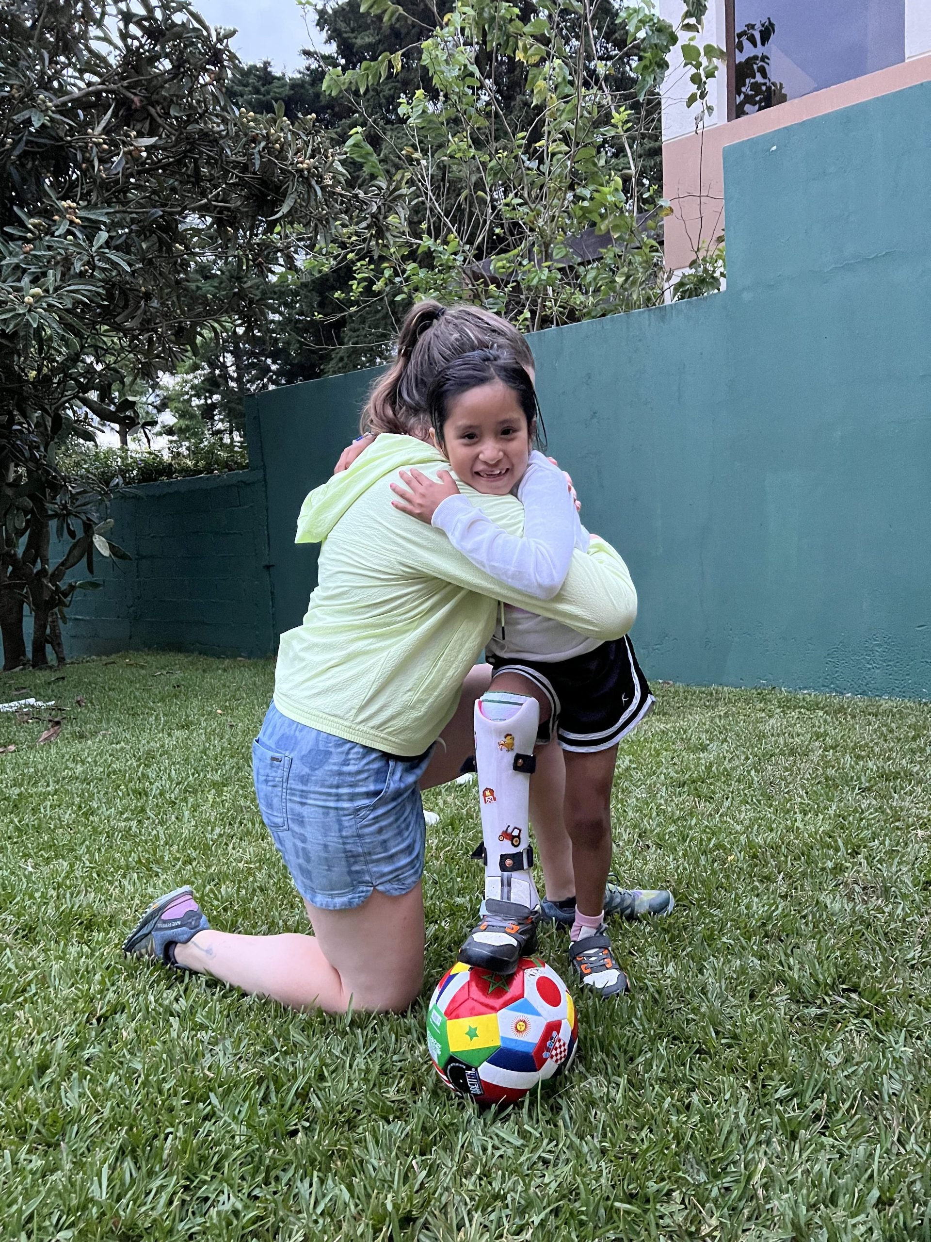 A woman hugging a young girl wearing a prosthetic leg standing with her foot on a soccer ball.