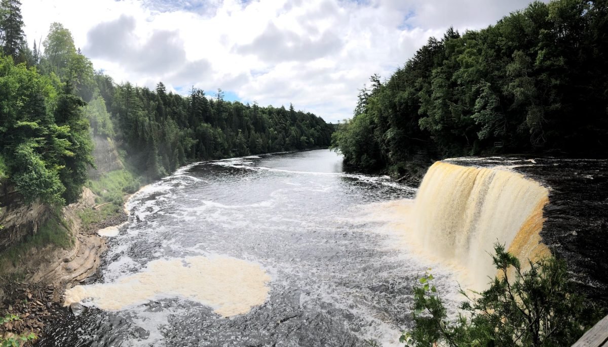 A waterfall with a light brown hue falling into a body of water below, and surrounded by forested banks on either side