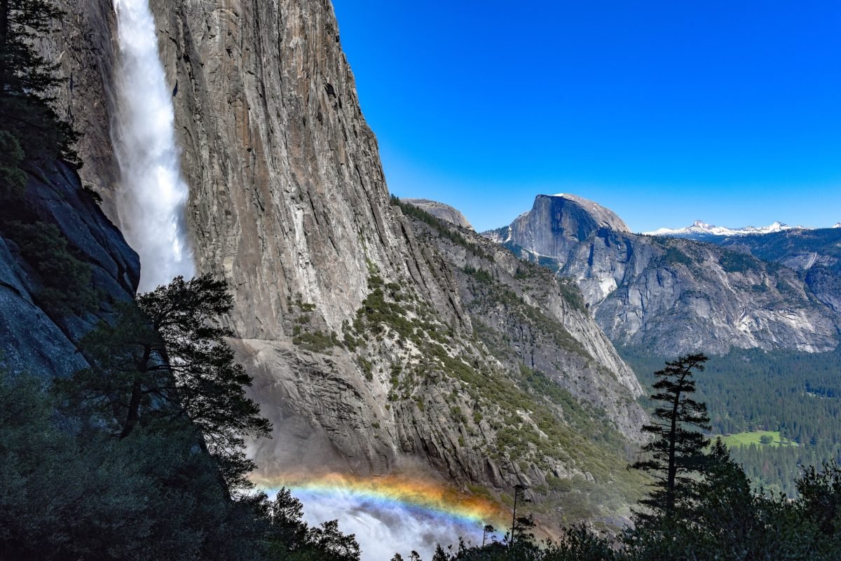 A landscape view of Half Dome from a waterfall lookout, with a rainbow in the mist of the falls.