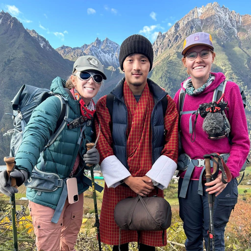 Two hikers and a guide smile at the camera in front of a mountainous landscape.