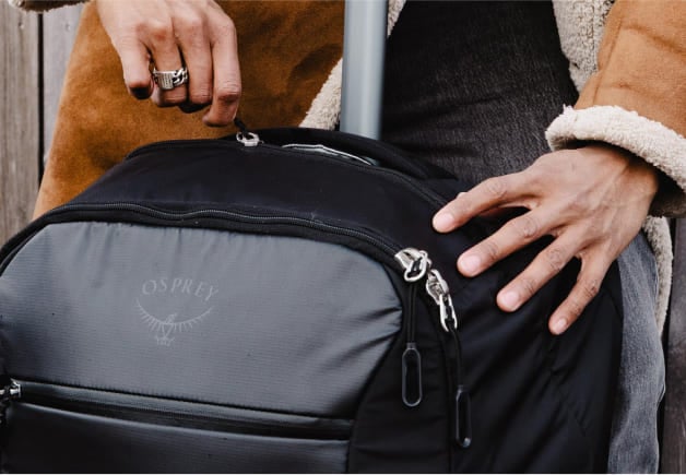 Close-up of a person’s hands holding the zippers of a black Osprey travel bag with a visible brand logo.