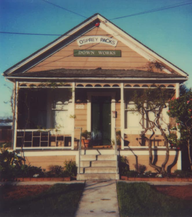 Faded, vintage photo of a peach-taupe Craftsman-style house with a front porch, converted into a storefront with two signs that say, &ldquo;Osprey Packs&rdquo; and &ldquo;Down Works.&rdquo;