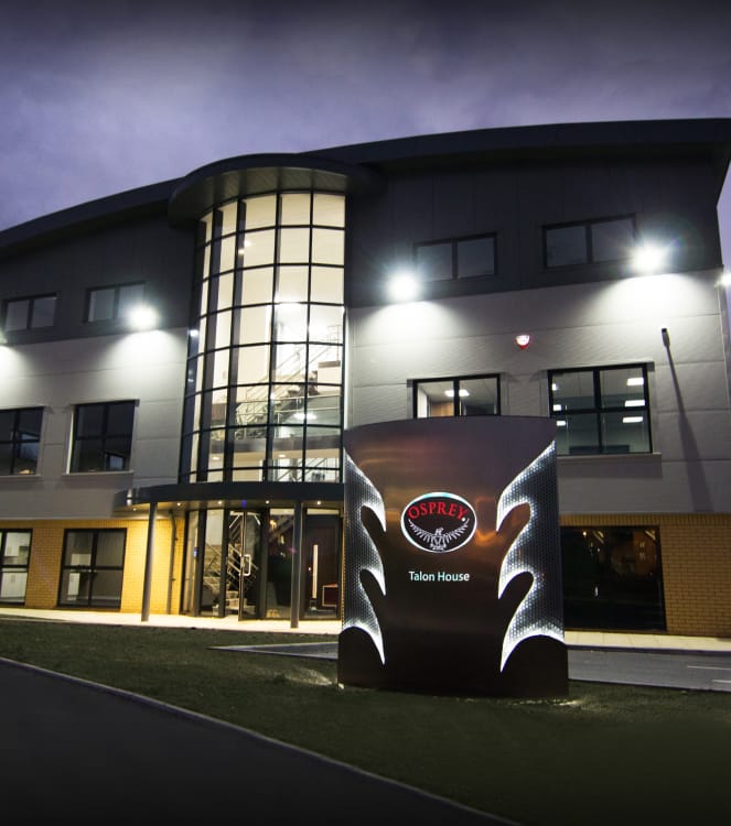 A nighttime photo of a 3-story office building with a curved roof and a multi-story glass atrium at the front entrance. There is a large, illuminated sign on a patch of grass in front of the building, featuring the Osprey logo, wing-like cutouts that are lit up, and the name &ldquo;Talon House.&rdquo;