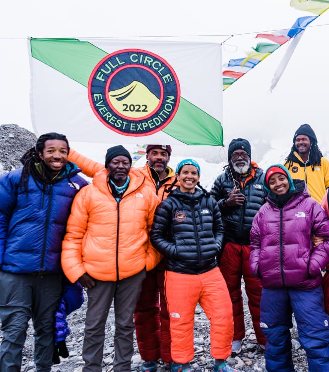 A team of seven climbers wearing
mountaineering pants and puffy coats under a white/green striped flag with a blue, red and yellow insignia that reads, &ldquo;Full Circle Everest Expedition 2022&rdquo; and Tibetan prayer flags blowing in the wind behind them.